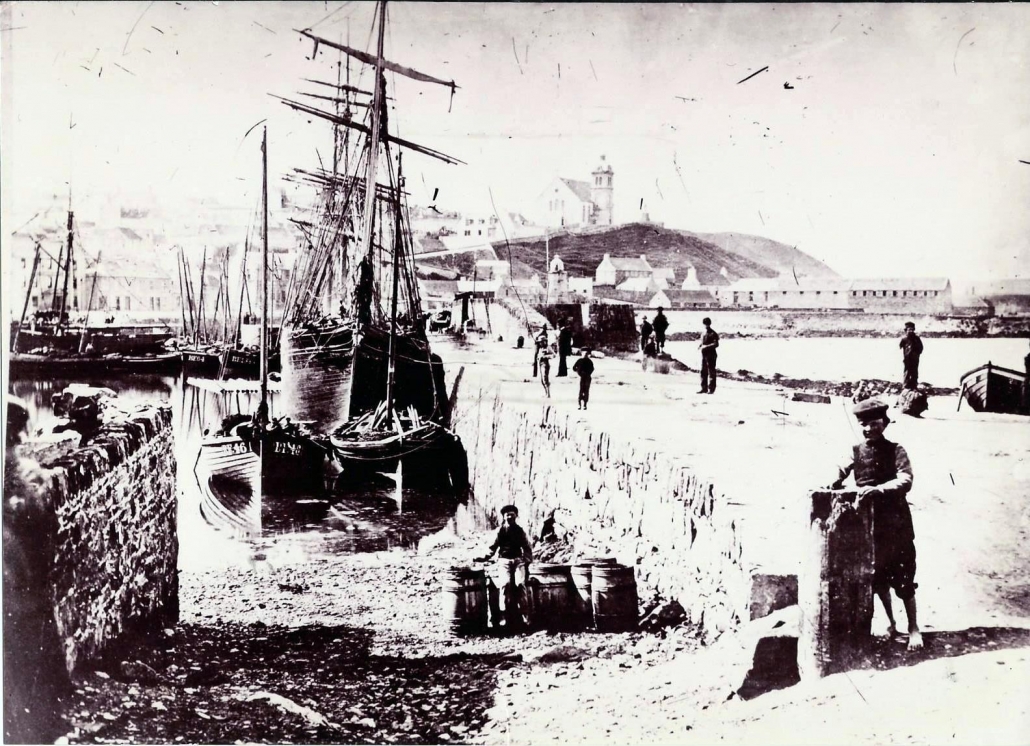 Black and white image showing a large sailing ship, various wooden fishing boats and Macduff church in the background.